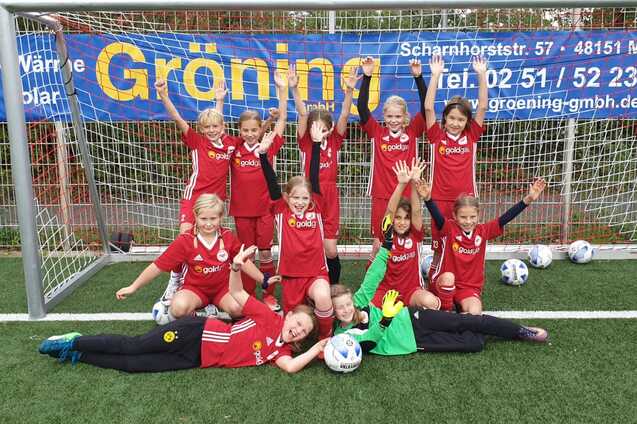 Gruppe von M&auml;dchen in roten Fu&szlig;balltrikots posiert fr&ouml;hlich vor einem Fu&szlig;balltor, umgeben von Fu&szlig;b&auml;llen.