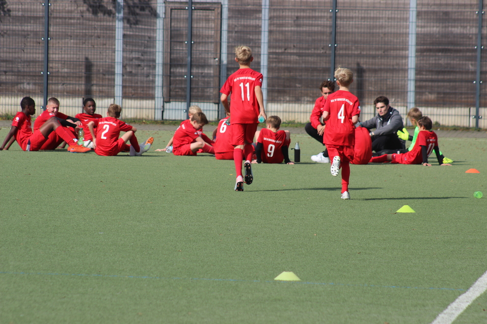 Zwei Kinder in roten Fu&szlig;balltrikots laufen auf einem Sportplatz, w&auml;hrend andere auf der Bank sitzen.