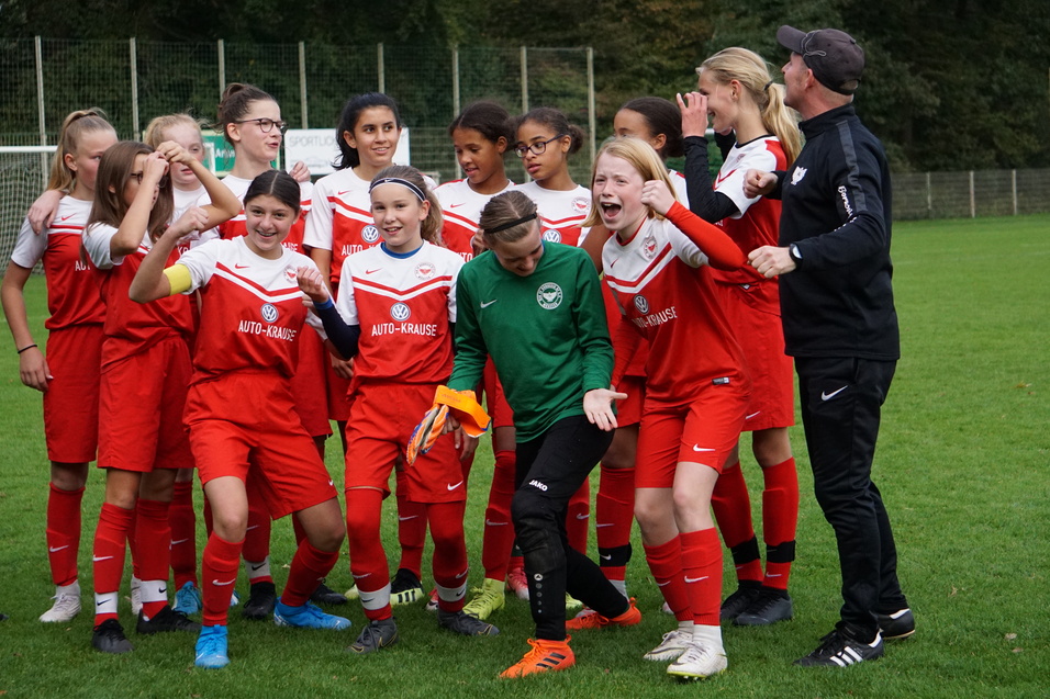 Junge Fu&szlig;ballmannschaft in roten Trikots steht lachend auf dem Rasen, einige Spielerinnen jubeln und posieren.