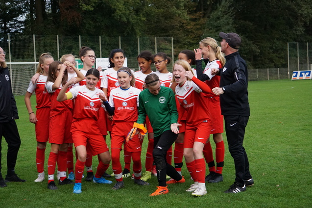 Junge Fu&szlig;ballmannschaft in roten Trikots steht lachend auf dem Rasen, einige Spielerinnen jubeln und posieren.