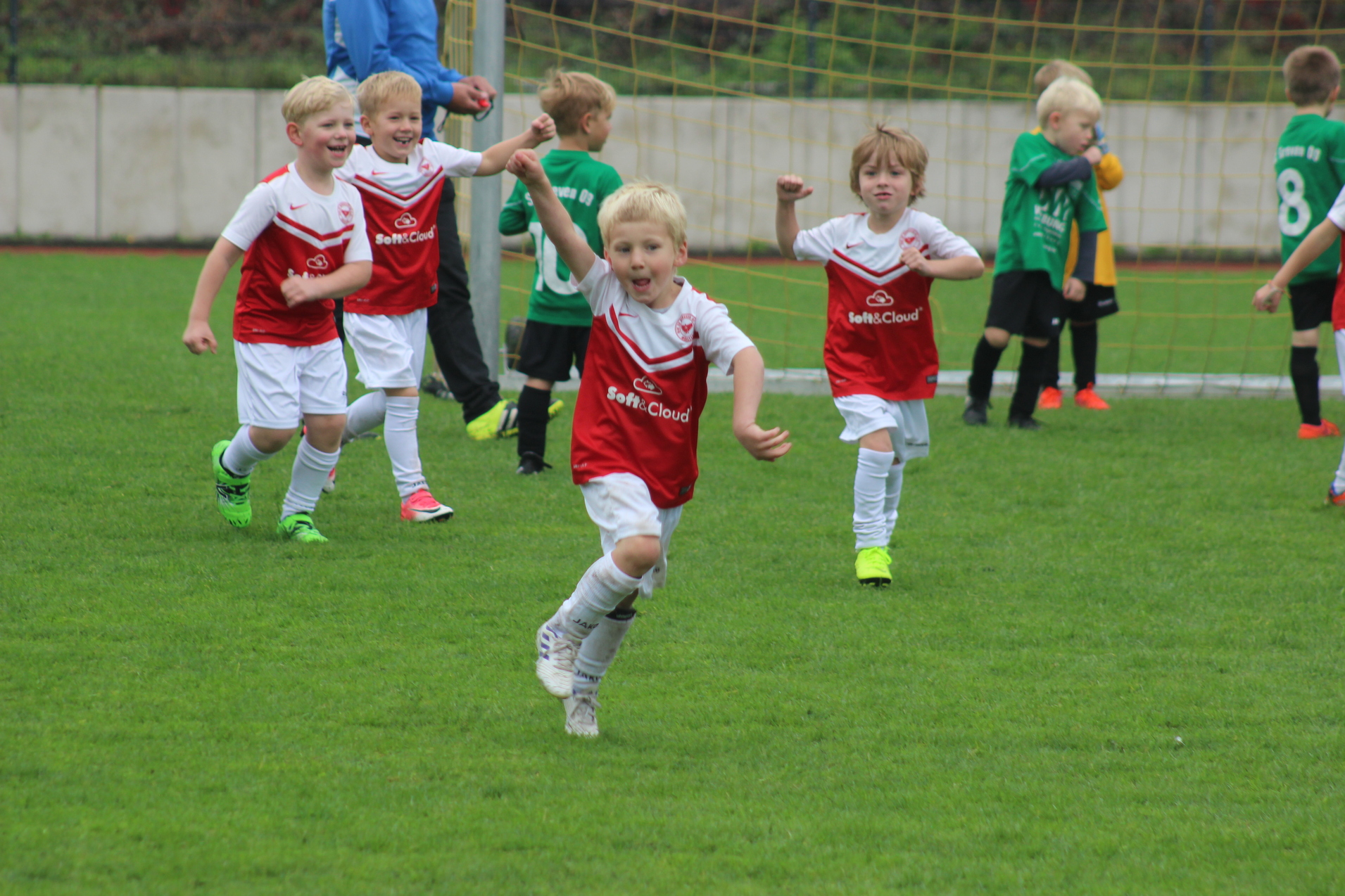 Ein jubelnder Junge in rotem Fu&szlig;balltrikot l&auml;uft &uuml;ber das Spielfeld, umgeben von weiteren spielenden Kindern.