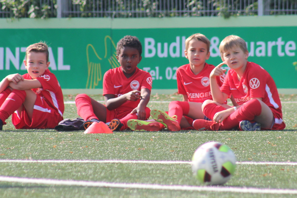 Vier Kinder in roten Fu&szlig;balltrikots sitzen auf einem Kunstrasenfeld, ein Fu&szlig;ball liegt vor ihnen.