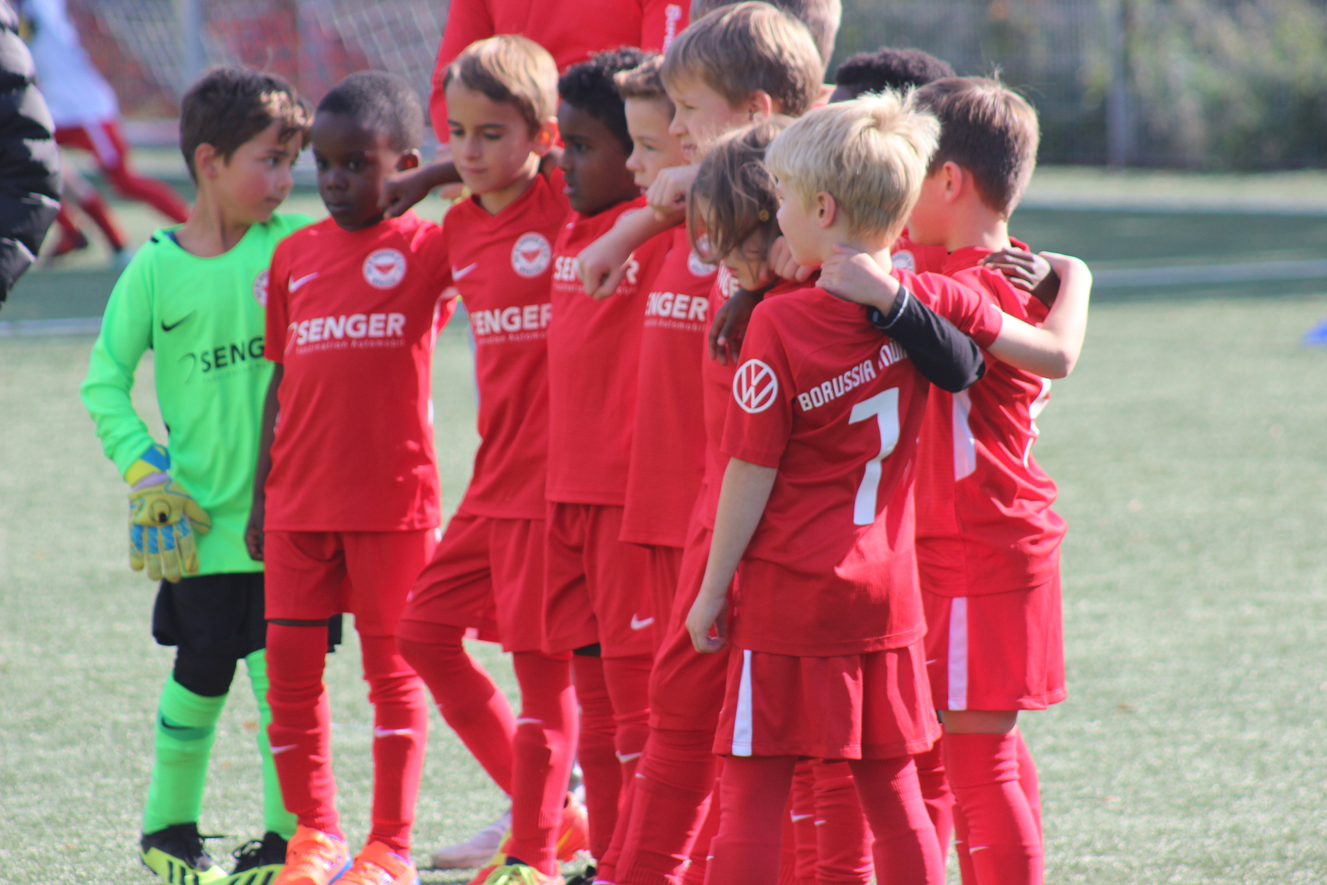 Gruppe von acht jungen Fu&szlig;ballspielern in roten Trikots, umarmt auf dem Spielfeld, mit einem Torwart in gr&uuml;n.