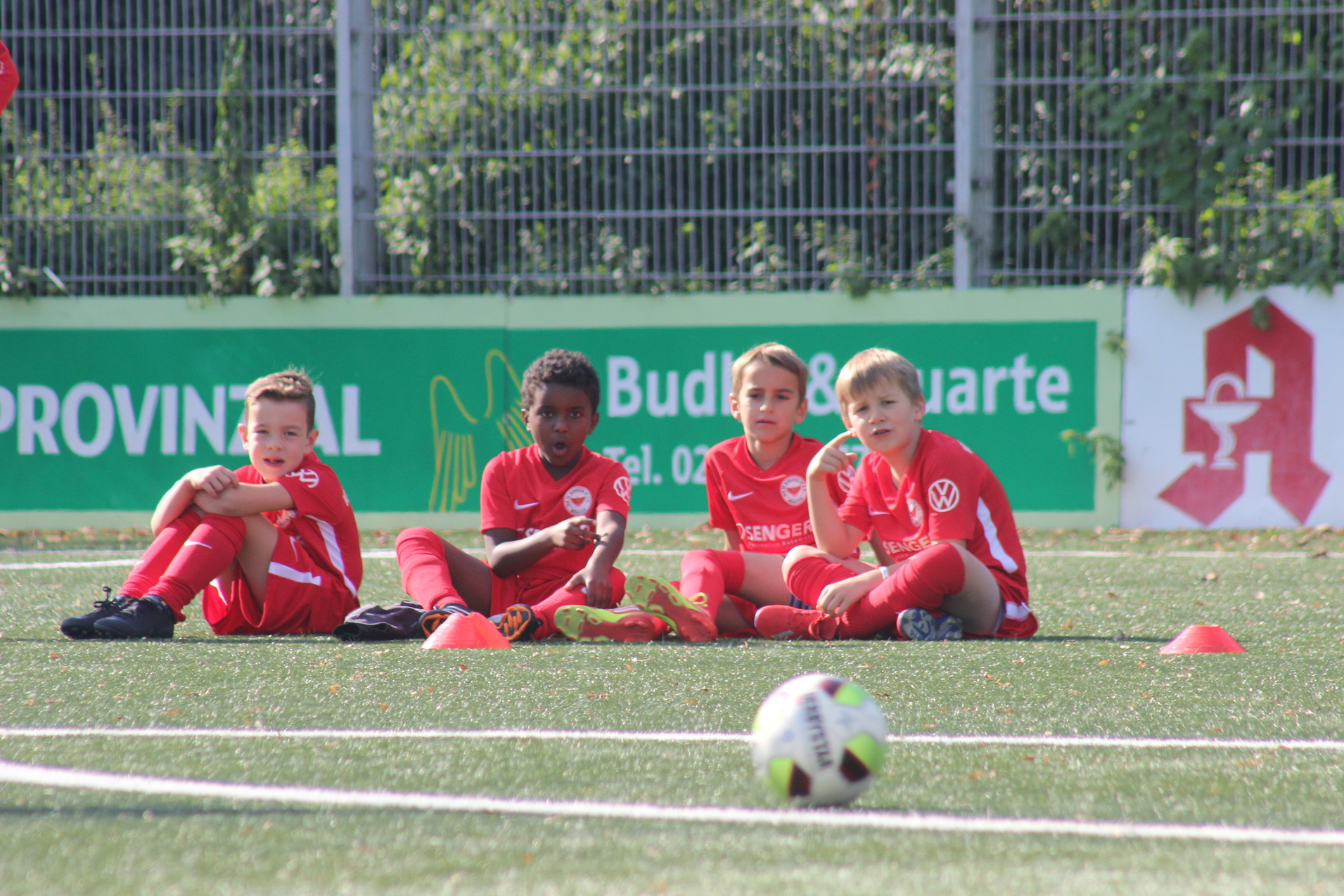 Vier Kinder in roten Fu&szlig;balltrikots sitzen auf einem Kunstrasenfeld, ein Fu&szlig;ball liegt vor ihnen.