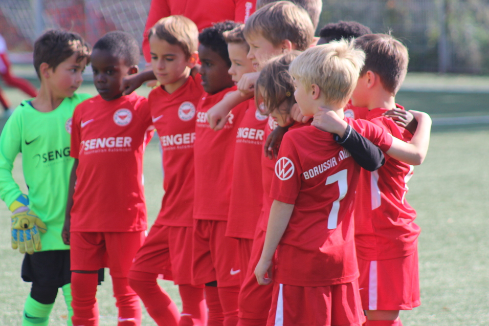 Gruppe von acht jungen Fu&szlig;ballspielern in roten Trikots, umarmt auf dem Spielfeld, mit einem Torwart in gr&uuml;n.