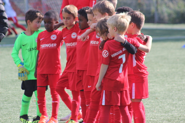 Gruppe von acht jungen Fu&szlig;ballspielern in roten Trikots, umarmt auf dem Spielfeld, mit einem Torwart in gr&uuml;n.
