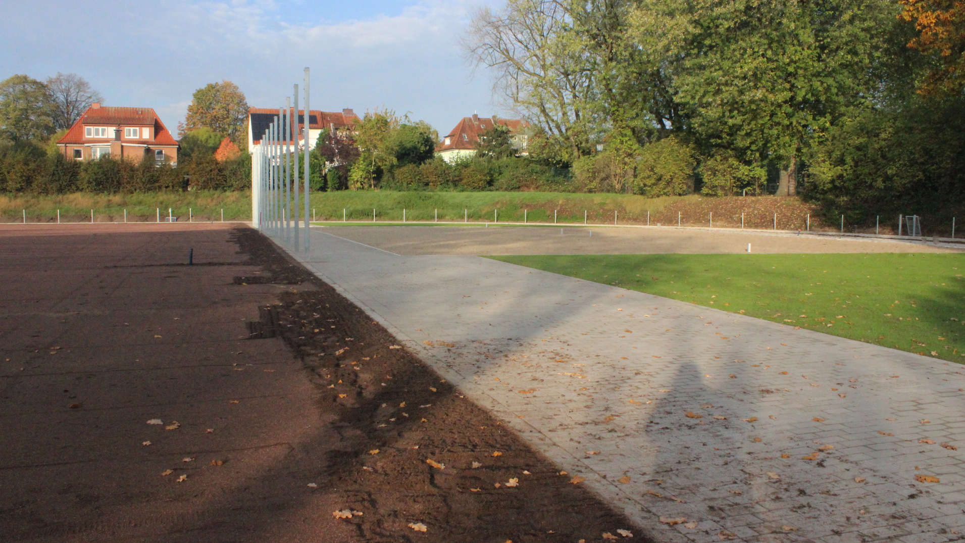 Blick auf einen neuen Sportplatz mit Asphalt- und Rasenbereichen sowie umliegenden B&auml;umen und H&auml;usern im Hintergrund.