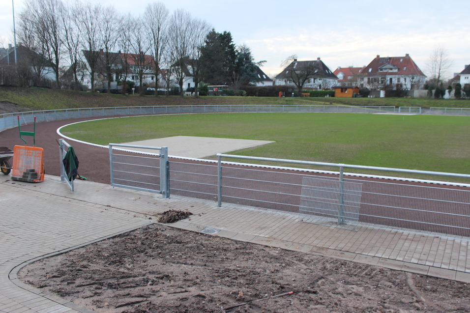 Blick auf einen Sportplatz mit Rasen, Laufbahn und einem Werkzeugbereich im Vordergrund. Wohnh&auml;user im Hintergrund.