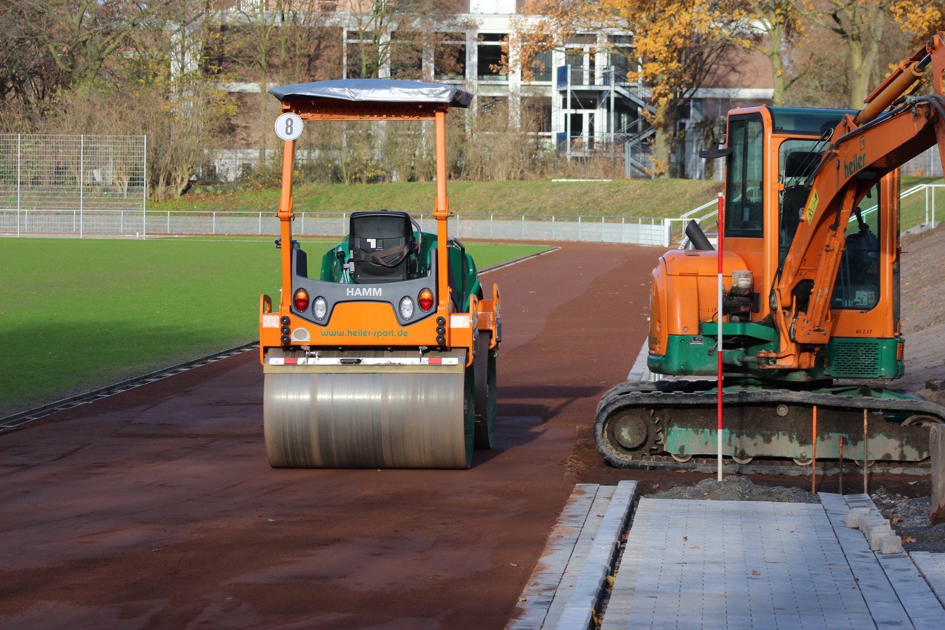 Bodenverdichter und Bagger auf einer Sportplatzbaufl&auml;che neben einer frisch verlegten Pflastersteinreihe.