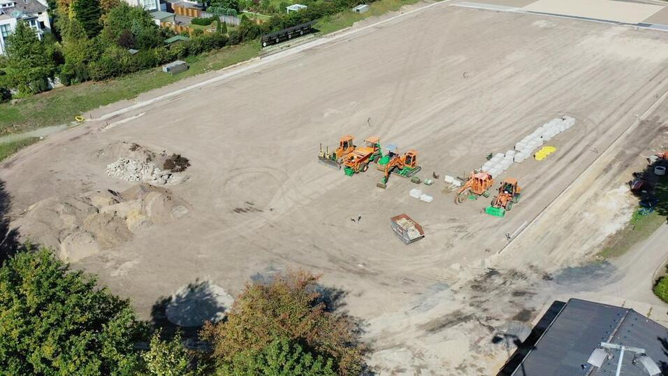 Blick auf eine gro&szlig;e, leere Baustelle mit Baumaschinen und umgebenden B&auml;umen und Wohngeb&auml;uden.