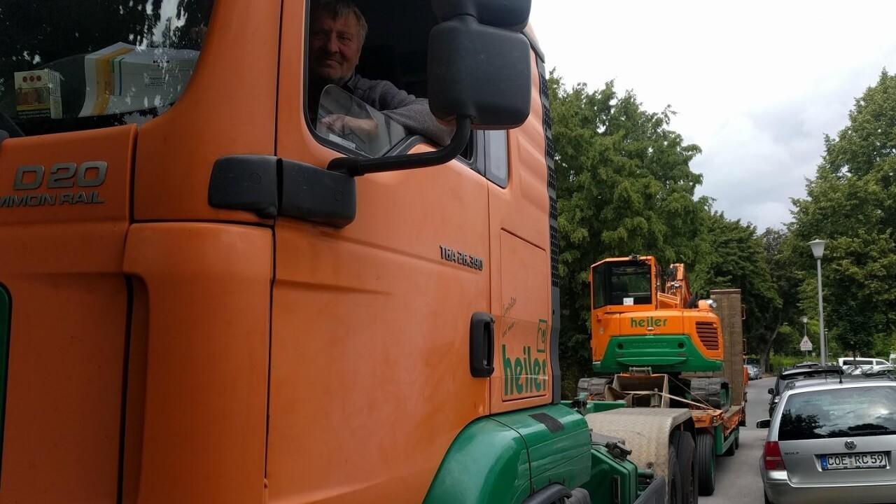 Lkw mit orangefarbener Kabine und Fahrer auf einer Stra&szlig;e, im Hintergrund eine orange Baustellenmaschine.