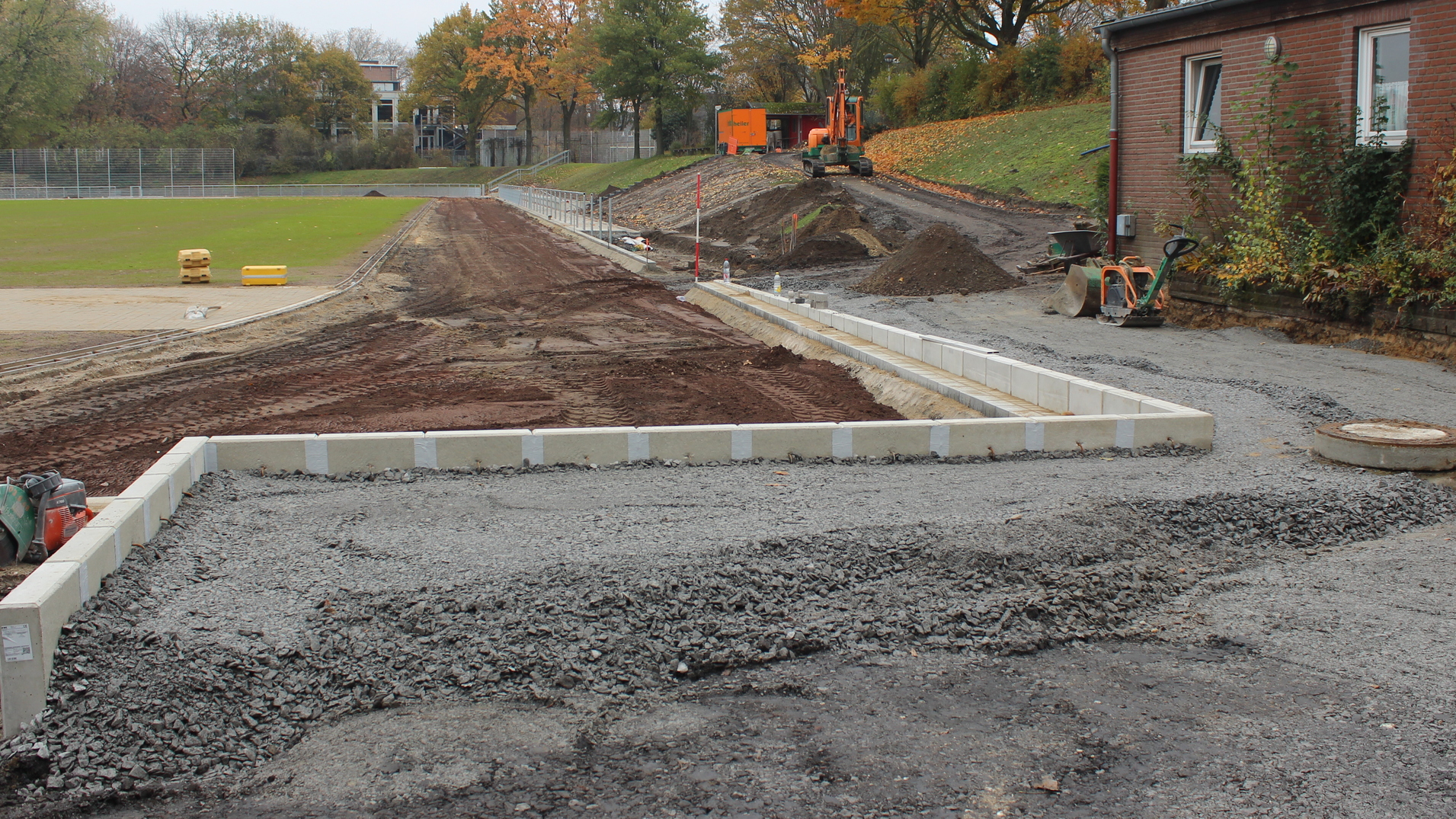 Bauarbeiten an einem Sportplatz: Fundamente aus Betonsteinen auf einem schottrigen Untergrund hinter einem Geb&auml;ude.