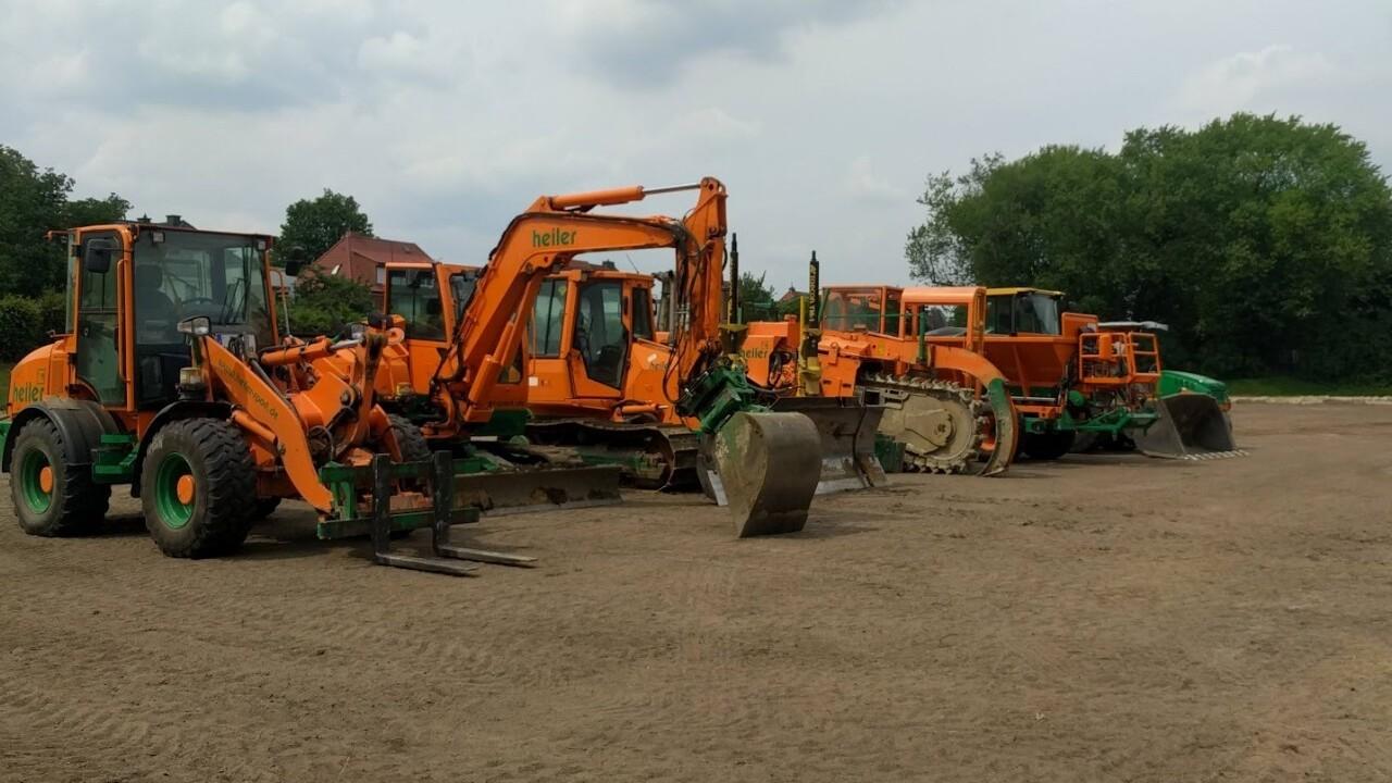 Reihe orangefarbener Baumaschinen auf einer unbefestigten Fl&auml;che unter einem wolkigen Himmel.