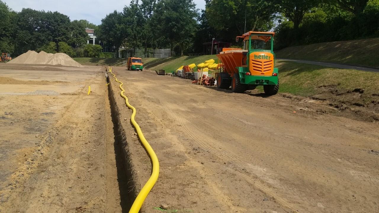 Baustelle mit gelbem Schlauch, Maschinen und Erde; im Hintergrund B&auml;ume und unebene Fl&auml;che f&uuml;r Arbeiten.