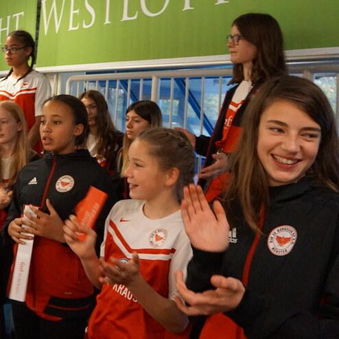 Mädchen in Sportkleidung stehen lächelnd zusammen, applaudieren und feuern eine Veranstaltung im Hintergrund an.