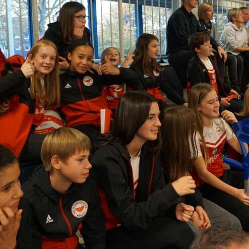 Gruppe von Jugendlichen in Sportkleidung, die auf Tribüne einer Veranstaltung sitzen und lächeln sowie jubeln.