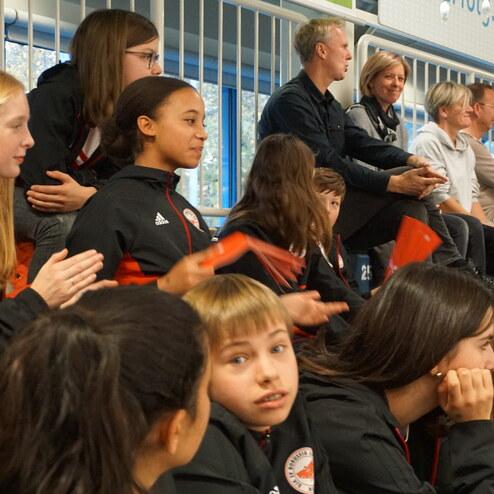 Gruppe von Jugendlichen in Sportjacken sitzt auf Tribüne, einige applaudieren, während Erwachsene im Hintergrund zuschauen.