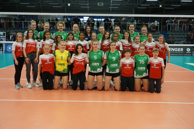 Gruppenfoto von zwei Volleyballmannschaften mit Spielerinnen in roten und gr&uuml;nen Trikots auf einem Volleyballfeld.