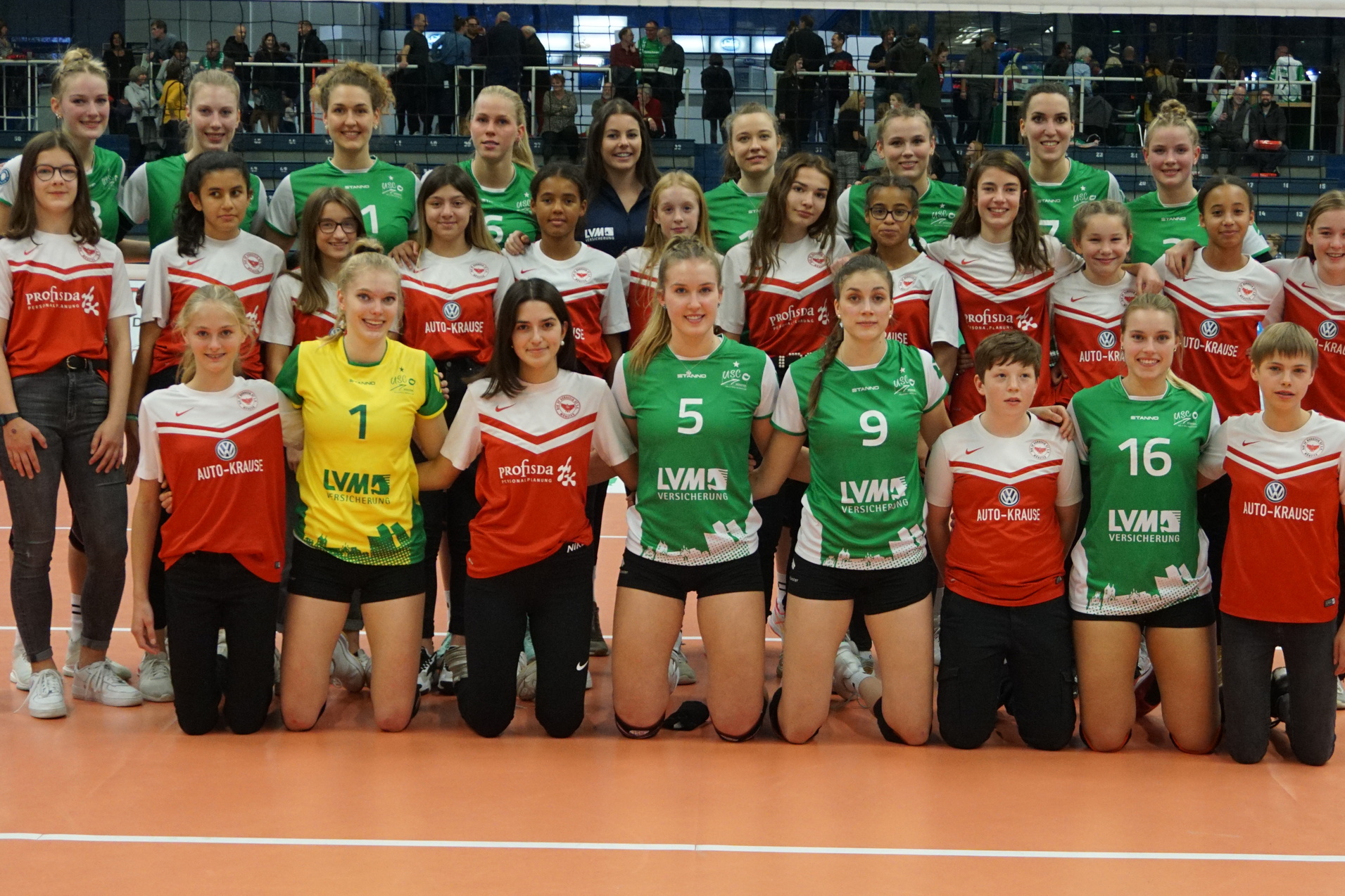 Gruppenfoto von zwei Volleyballmannschaften mit Spielerinnen in roten und gr&uuml;nen Trikots auf einem Volleyballfeld.