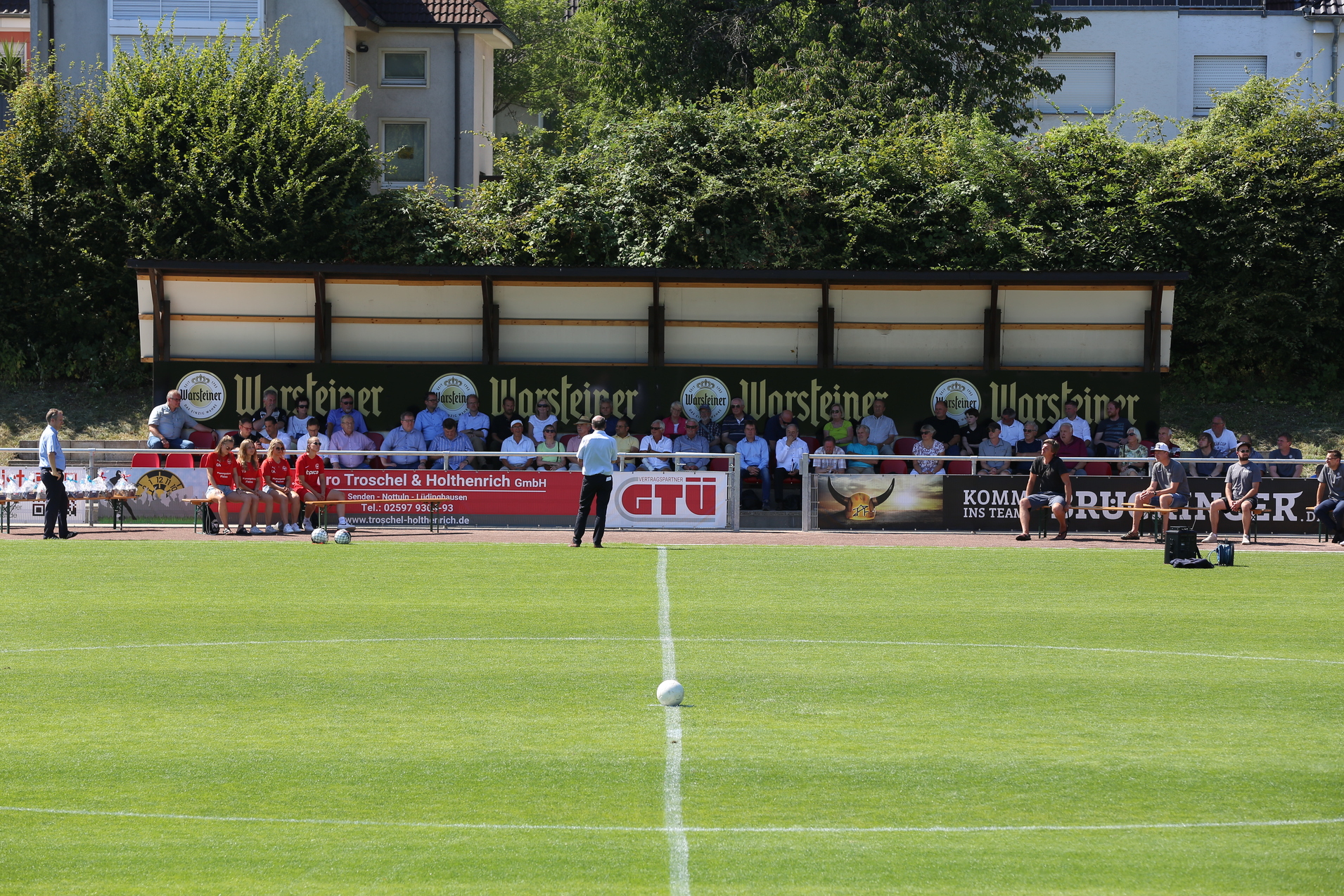 Blick auf ein Fu&szlig;ballfeld mit einer Zuschauertrib&uuml;ne und mehreren Menschen auf der Bank, bei sonnigem Wetter.