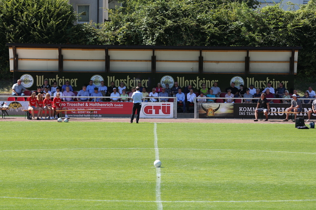 Blick auf ein Fu&szlig;ballfeld mit einer Zuschauertrib&uuml;ne und mehreren Menschen auf der Bank, bei sonnigem Wetter.