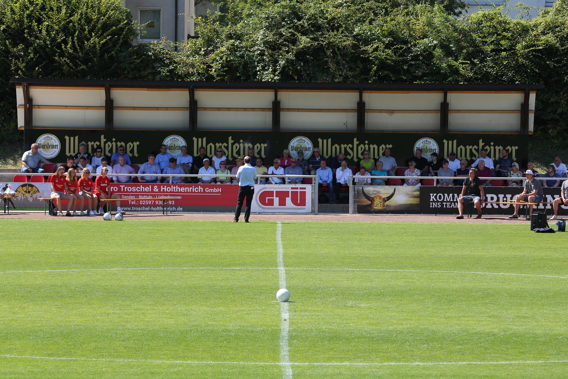 Blick auf ein Fu&szlig;ballfeld mit einer Zuschauertrib&uuml;ne und mehreren Menschen auf der Bank, bei sonnigem Wetter.