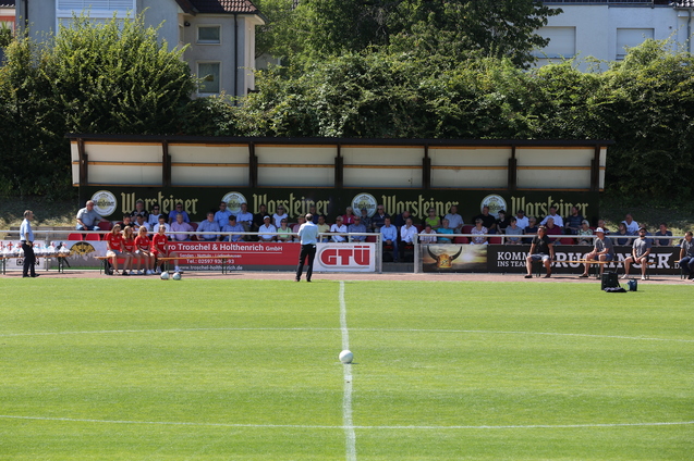 Blick auf ein Fu&szlig;ballfeld mit einer Zuschauertrib&uuml;ne und mehreren Menschen auf der Bank, bei sonnigem Wetter.
