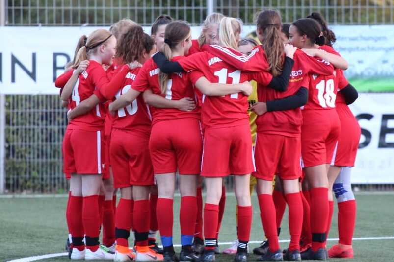 M&auml;dchenfu&szlig;ballmannschaft in roten Trikots steht eng umschlungen auf dem Spielfeld vor einem Werbebanner.