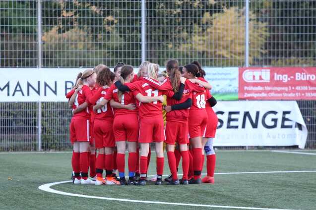 M&auml;dchenfu&szlig;ballmannschaft in roten Trikots steht eng umschlungen auf dem Spielfeld vor einem Werbebanner.