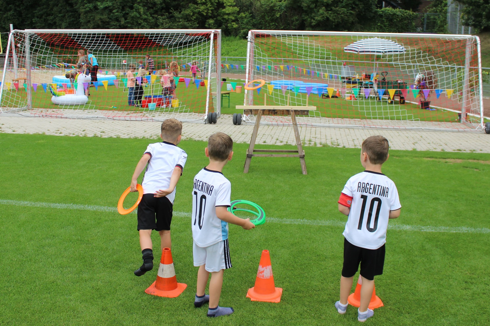 Drei Kinder werfen Reifen auf einem Sportplatz, im Hintergrund findet eine Feier mit Wasserspielen statt.