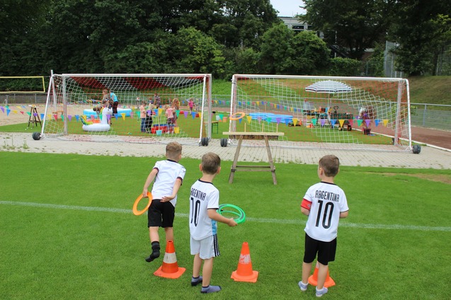 Drei Kinder werfen Reifen auf einem Sportplatz, im Hintergrund findet eine Feier mit Wasserspielen statt.