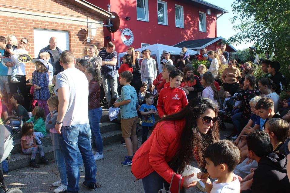 Gruppe von Kindern und Erwachsenen versammelt sich vor einem roten Geb&auml;ude bei einer Veranstaltung im Freien.