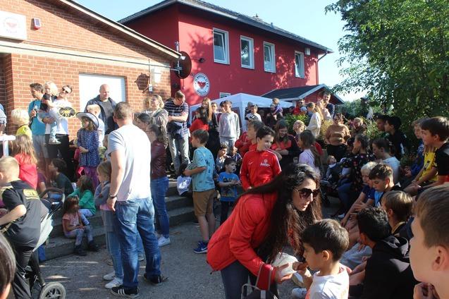 Gruppe von Kindern und Erwachsenen versammelt sich vor einem roten Geb&auml;ude bei einer Veranstaltung im Freien.
