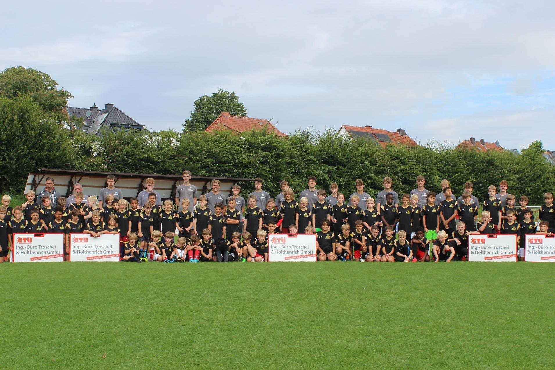 Gruppenfoto von Jugendlichen und Erwachsenen in Sporttrikots auf einem Fußballplatz, mit Mannschaftskästen im Hintergrund.
