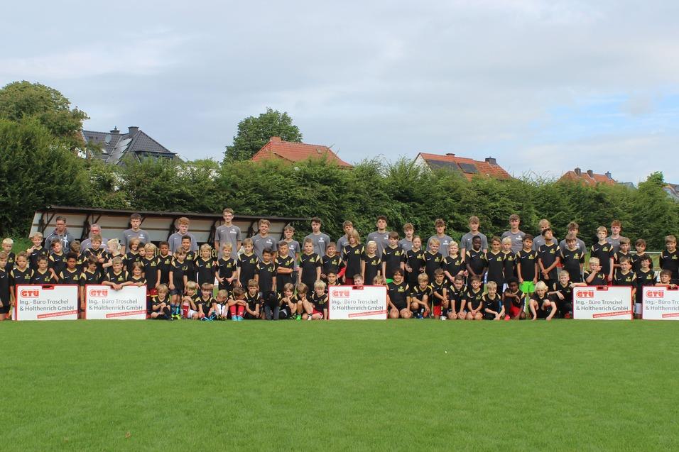 Gruppenfoto von Jugendlichen und Erwachsenen in Sporttrikots auf einem Fu&szlig;ballplatz, mit Mannschaftsk&auml;sten im Hintergrund.