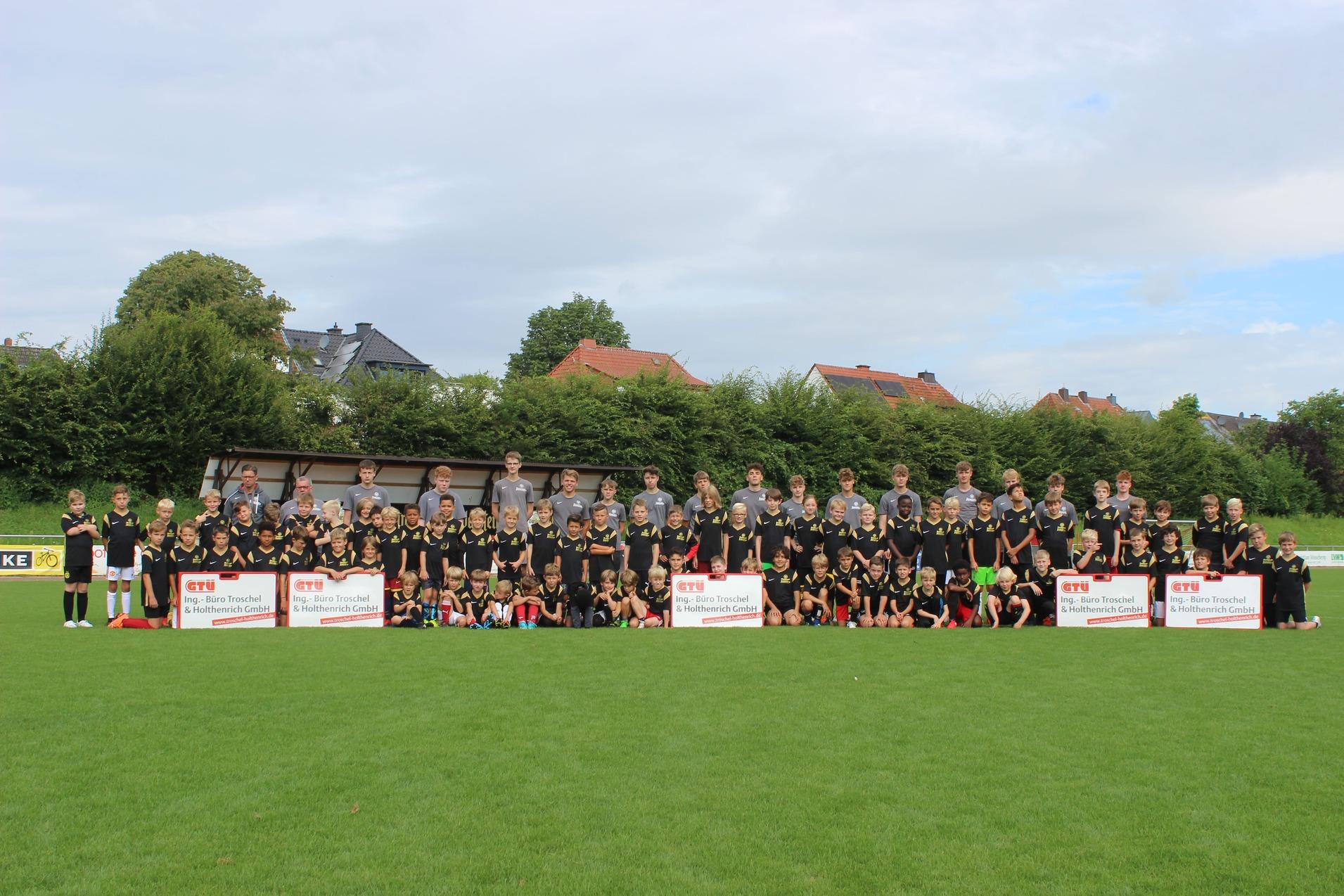 Gruppenfoto von Jugendlichen und Erwachsenen in Sporttrikots auf einem Fu&szlig;ballplatz, mit Mannschaftsk&auml;sten im Hintergrund.