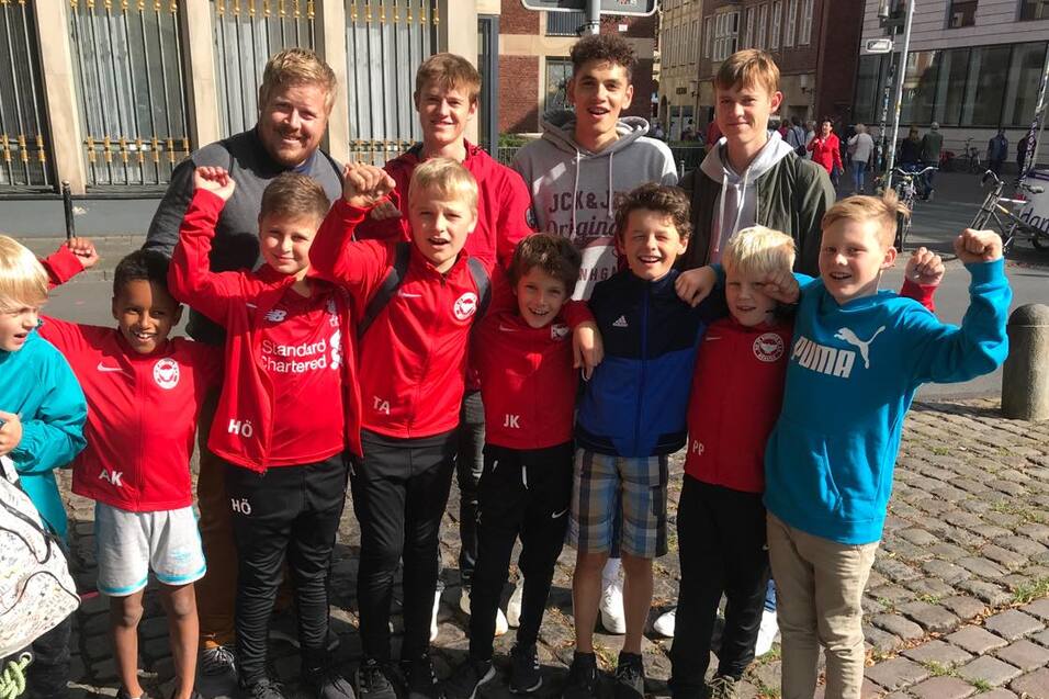 Gruppe von zehn Jungen in roten Trikots und Sportkleidung, l&auml;chelnd auf einem Kopfsteinpflasterplatz versammelt.