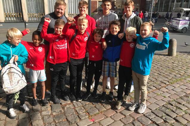Gruppe von zehn Jungen in roten Trikots und Sportkleidung, l&auml;chelnd auf einem Kopfsteinpflasterplatz versammelt.
