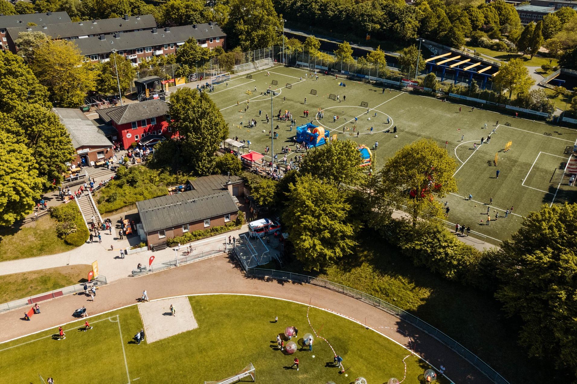 Luftaufnahme eines Sportplatzes mit Fu&szlig;ballspielern, bunten Zelten und umgebenden B&auml;umen in einem st&auml;dtischen Umfeld.