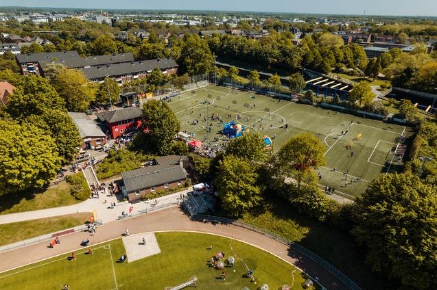 Luftaufnahme eines Sportplatzes mit Fu&szlig;ballspielern, bunten Zelten und umgebenden B&auml;umen in einem st&auml;dtischen Umfeld.