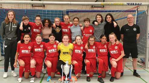 Mädchenfußballmannschaft in roten Trikots posiert mit Pokal am Hallenfußballtor nach einem erfolgreichen Spiel.