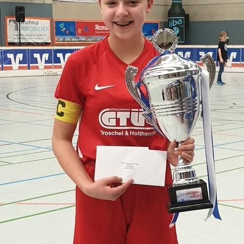 Mädchen in rotem Sporttrikot hält einen Pokal und eine Urkunde in einer Sporthalle.