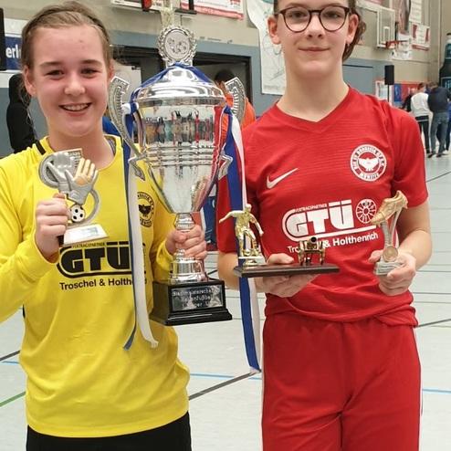 Zwei junge Fußballspielerinnen halten Trophäen, eine in gelber und die andere in roter Sportkleidung, nach einem Turnier.