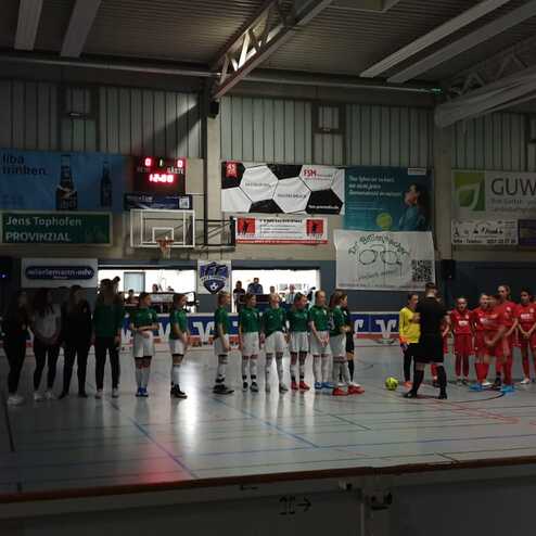 Zwei Fußballmannschaften in einem Sporthalle stehen sich gegenüber, bereit für das Spiel. Plakatwände im Hintergrund.