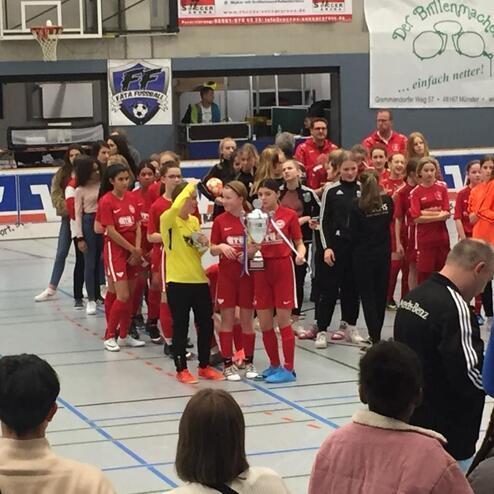 Mädchenfußballmannschaft in roten Trikots nimmt Pokal entgegen, Zuschauer im Hintergrund verfolgen die Ehrung.