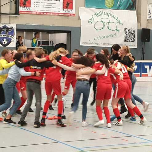 Gruppe junger Frauen in roten Trikots umarmt sich jubelnd in einer Sporthalle nach einem Spiel.