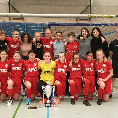 Mädchenfußballmannschaft in roten Trikots posiert mit Pokal am Hallenfußballtor nach einem erfolgreichen Spiel.