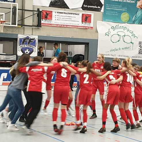 Mädchenfußballteam in roten Trikots umarmt sich in einer Sporthalle vor einer unerkennbaren Zuschauertribüne.