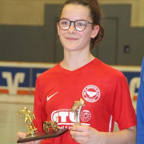 Mädchen in rotem Fußballtrikot hält mehrere Pokale in der Sporthalle. Sie trägt eine Brille und lächelt.