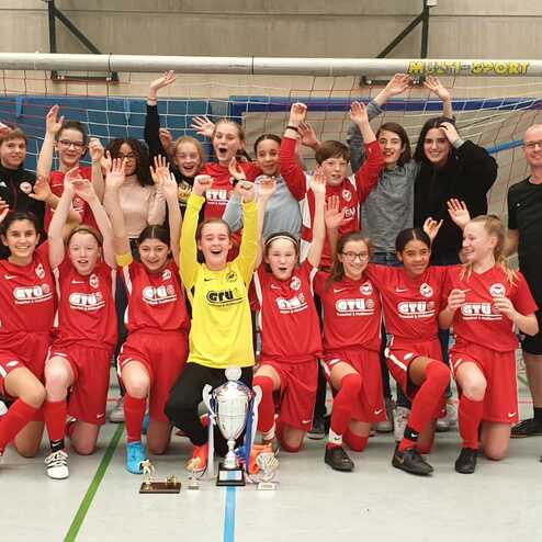 Mannschaftsbild einer Fußballjugend in roten Trikots mit Pokal, jubelnd vor einem Tor in einer Sporthalle.